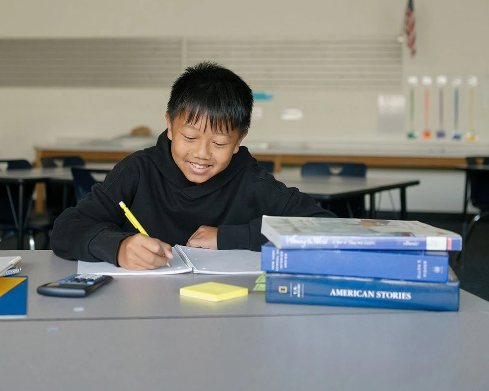 A student writes in a notebook at a classroom desk.