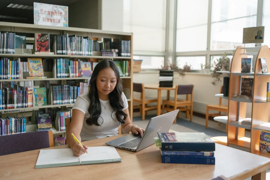 A student studies with a notebook and laptop in a library.